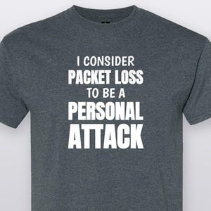 May include: A dark heather gray short-sleeved t-shirt featuring the white text I CONSIDER PACKET LOSS TO BE A PERSONAL ATTACK printed in a bold, sans-serif font across the center of the chest. The garment has a crew neckline and a standard fit.