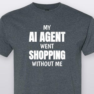 May include: A dark heather gray short-sleeved t-shirt featuring the white text MY AI AGENT WENT SHOPPING WITHOUT ME printed in a bold, sans-serif font across the center of the chest. The garment has a crew neckline and a standard fit.