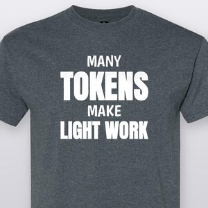 May include: A dark heather gray short-sleeve t-shirt featuring the white text "MANY TOKENS MAKE LIGHT WORK" printed in a bold, sans-serif font across the chest. The garment has a crew neckline and a standard fit with a visible heathered texture.