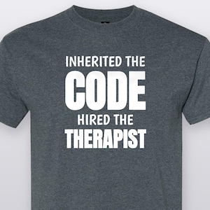 May include: A dark heather gray short-sleeved t-shirt featuring white text printed on the chest that reads INHERITED THE CODE HIRED THE THERAPIST. The shirt has a crew neckline and a standard fit, constructed from a soft, heathered fabric blend.