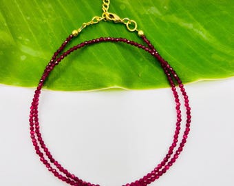 May include: A delicate, double-strand necklace featuring small, round, deep red beads. The necklace has a gold-colored chain and clasp. The jewelry is displayed against a bright green leaf and a white background.