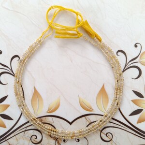 Peut inclure: Un collier de perles de pierres précieuses brun clair avec un pompon jaune. Les perles sont enfilées sur un cordon fin et sont disposées en cercle.