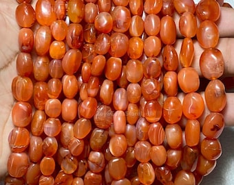 Puede incluir: Un conjunto de cuentas de cornalina pulidas, de forma ovalada, en tonos naranja y marrón rojizo. Las cuentas están ensartadas, mostrando sus variaciones naturales y translucidez. Las cuentas tienen un acabado liso y brillante, de tamaños variados.