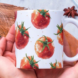May include: White ceramic mug with a repeating pattern of red tomatoes on a white background. The mug is being held by a person's hands.