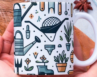 May include: White ceramic mug with a colorful pattern of gardening tools and plants. The mug is being held by a person's hands.