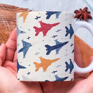 May include: White ceramic mug featuring a repeating pattern of colorful fighter jets in red, blue, and orange on a cream background. The mug has a curved handle and is held in both hands.