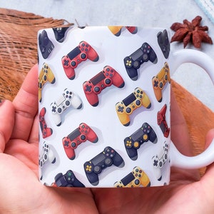 May include: White ceramic mug featuring a pattern of colourful video game controllers. The mug is being held by a person's hands.