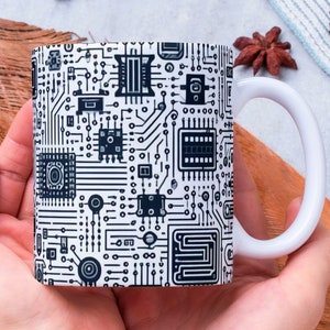 Puede incluir: Una taza de cerámica blanca con un diseño de placa de circuito negro y blanco. La taza está siendo sostenida en dos manos.