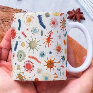 Puede incluir: Una taza de cerámica blanca con un patrón colorido de varias bacterias y microorganismos. La taza está siendo sostenida por las manos de una persona.