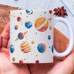 May include: A white ceramic mug with a colorful space-themed design featuring planets, stars, and a galaxy print. The mug is being held by a person's hands.