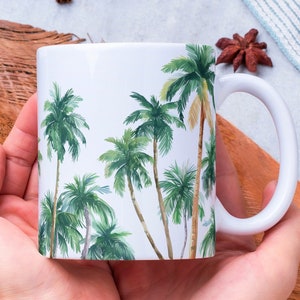 May include: White ceramic mug with a tropical palm tree design. The mug is being held by a person wearing a white sweater.