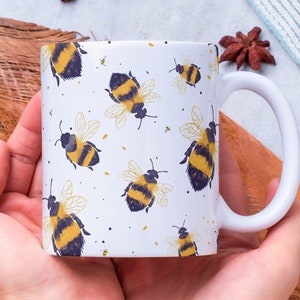 May include: White ceramic mug with a pattern of yellow and black bumblebees. The mug is being held by a person's hands.
