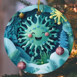 Peut inclure: Une boule de Noël ronde avec un motif découpé en papier bleu, vert et blanc. Un virus vert souriant avec un visage blanc est au centre de l'ornement. La boule a une ficelle dorée pour l'accrocher.