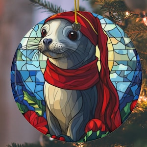 Peut inclure: Une boule de Noël ronde représentant un phoque de dessin animé portant une écharpe rouge et un bonnet de Père Noël rouge. La boule a un design en vitrail avec des couleurs bleues, rouges et vertes.