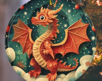 May include: A round Christmas ornament featuring a cartoon dragon with orange scales and wings. The dragon is set against a backdrop of a snowy forest with a teal background. The ornament hangs from a gold string.
