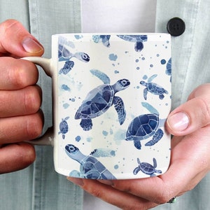 Può includere: Una tazza in ceramica bianca con un motivo di tartarughe marine in acquerello blu. La tazza ha un manico ed è tenuta da una persona.