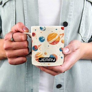 May include: White ceramic mug with a space-themed design featuring planets, stars, and a galaxy background. The mug has a black handle and the name "JERRY" printed on the bottom.