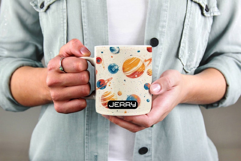 May include: White ceramic mug with a space-themed design featuring planets, stars, and a galaxy background. The mug has a black handle and the name "JERRY" printed on the bottom.