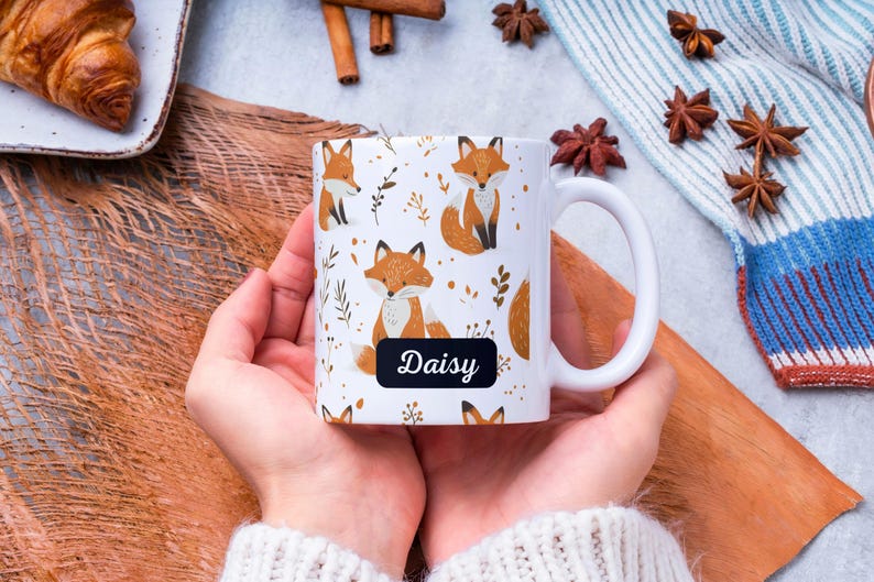 May include: White ceramic mug with a handle, featuring a repeating pattern of orange foxes and gold floral designs. The mug has a black label with the name "Daisy" written in white.