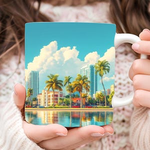 May include: White ceramic mug with a colorful image of a beach scene with palm trees and buildings. The sky is blue with white clouds. The water is blue and reflects the sky and buildings.