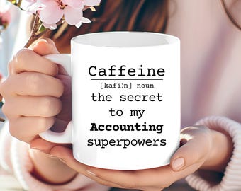 May include: White ceramic mug with the text "Caffeine [kafi:n] noun the secret to my Accounting superpowers". The mug is held by a person with pink flowers in the background.