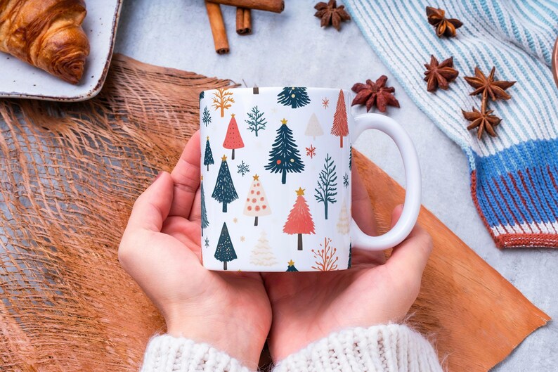 Puede incluir: Taza de cer&aacute;mica blanca con un patr&oacute;n de &aacute;rbol de Navidad. La taza est&aacute; decorada con &aacute;rboles rojos, verdes, azules y blancos, y estrellas doradas. La taza est&aacute; siendo sostenida por las manos de una persona.