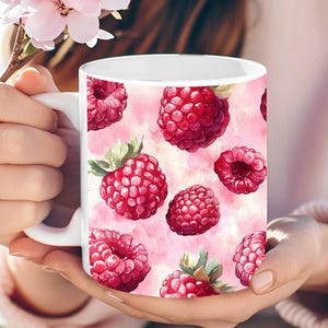 Peut inclure: Une tasse en céramique blanche avec un motif aquarelle rose et blanc représentant un motif répété de framboises rouges.