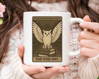 May include: White ceramic mug with an owl illustration, wings spread, on books. The design is framed with the text "THE WISE OWL". The background shows a library. The mug is held by a person.