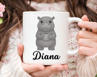 May include: White ceramic mug featuring a gray hippo holding a coffee cup. The hippo is illustrated in a cartoon style. The name "Diana" is written in a cursive font below the hippo. The mug has a curved handle.