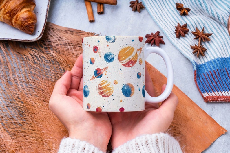 May include: A white ceramic mug with a colorful space-themed design featuring planets, stars, and a galaxy print. The mug is being held by a person's hands.