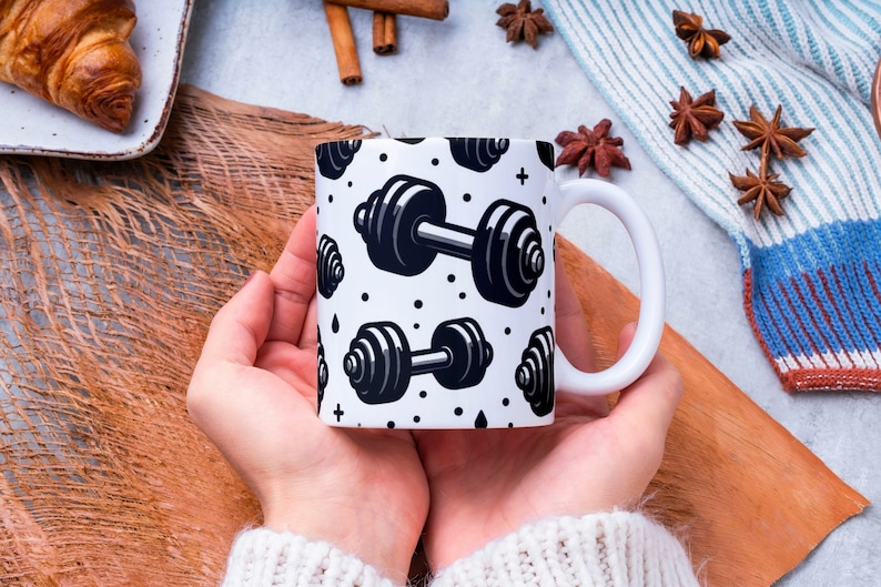 May include: White ceramic mug with a black and white pattern of dumbbells. The mug is being held by a person's hands.