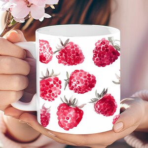 Op de afbeelding: Witte keramische mok met een patroon van aquarel frambozen in rood en roze. De bessen hebben groene bladeren en zijn verspreid over het oppervlak van de mok. De mok wordt in de handen van een persoon gehouden.