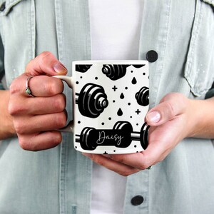 May include: White ceramic mug with a black and white pattern of dumbbells and the name "Daisy" printed on the front.