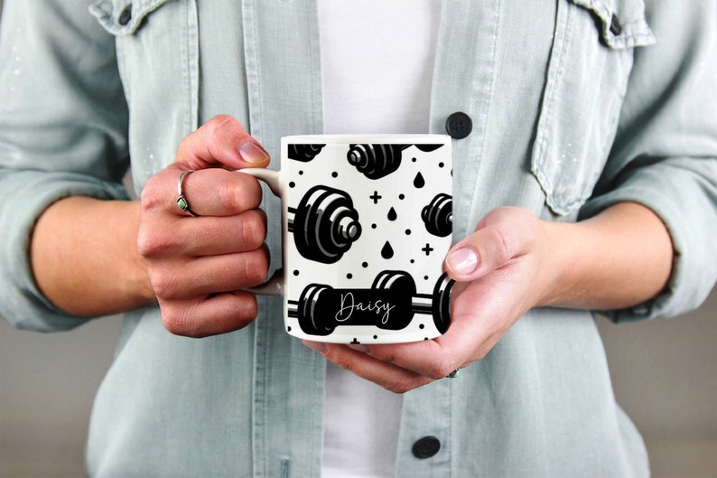 May include: White ceramic mug with a black and white pattern of dumbbells and the name "Daisy" printed on the front.