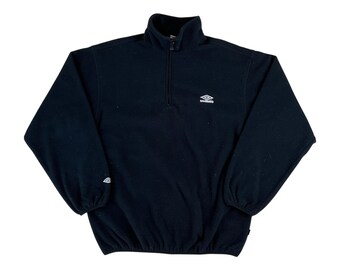 May include: A black long-sleeve fleece pullover featuring a quarter-zip mock neck collar, elasticized cuffs, and a cinched hem. The Umbro logo is embroidered in white on the left chest and a smaller logo is on the right sleeve cuff.