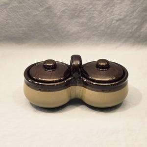 May include: A two-section ceramic condiment server with a handle. The server has a light tan base with a dark brown rim and lid. The lids have a small knob on top.