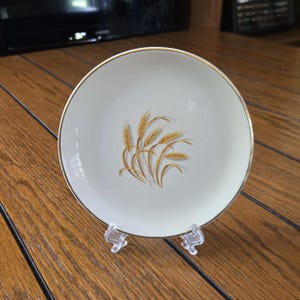 May include: A cream-colored plate with a gold rim and a wheat stalk design in shades of brown and gold. The plate is displayed on a clear plastic stand, showcasing its decorative elements. This vintage plate is a classic piece of tableware.