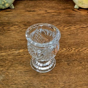May include: A clear, vintage glass votive candle holder with a detailed, cut-glass design. The candle holder has a round base, a short stem, and a wide, open top. The glass has a diamond and floral pattern. The votive holder is on a wooden surface.