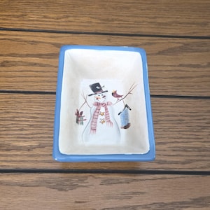 May include: A rectangular ceramic dish with a light blue border. The interior features a hand-painted snowman wearing a black top hat and a pink scarf. A bird and a birdhouse are also painted on the dish.