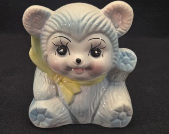 May include: A ceramic bear cub figurine with a light blue body, pink ears, and a yellow scarf. The bear has large eyes, rosy cheeks, and is holding a small blue object. The figurine is in a sitting position.