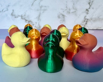 May include: A group of 12 colorful rubber duck toys in various shades of yellow, green, red, and brown. The ducks are arranged in a semi-circle on a white marble surface.