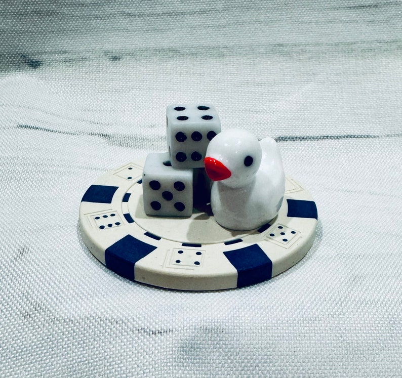 Puede incluir: Una figurita de pato de cer&aacute;mica blanca con un pico rojo se sienta en un tablero de juego circular azul y blanco con tres dados blancos con puntos negros.