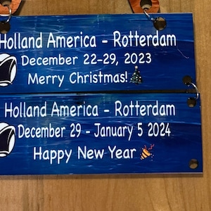 以下が含まれることがあります： 船のロゴと白い文字が書かれた青い木製の看板が2つ。上の看板には「Holland America - Rotterdam 2023年12月22日～29日 メリークリスマス！」と書いてあります。下の看板には「Holland America - Rotterdam 2024年1月5日 新年明けましておめでとうございます」と書いてあります