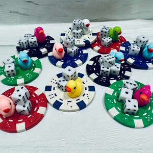 Puede incluir: Nueve fichas de casino de colores con dados y figuras de patos de goma en la parte superior. Cada ficha tiene un color y un dise&ntilde;o diferente.