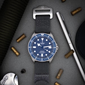 May include: A silver and blue Tudor Pelagos Chronometer watch with a black nylon strap. The watch has a blue dial with silver hands and markers. The watch is on a grey surface with a pen, bullets, and a roll of green fabric.