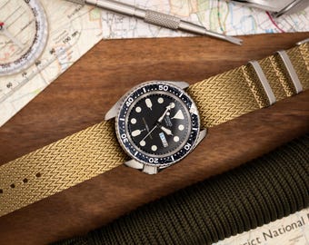 May include: A black and silver wristwatch with a black face and a gold nylon watch strap. The watch has a date window and a rotating bezel.