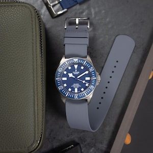May include: A silver wristwatch with a blue face and grey strap. The watch is shown with a spare blue strap and a green leather case. The watch face has white markings and the brand name.