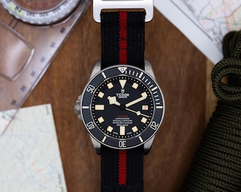 May include: A silver wristwatch with a black face and red accents on the black nylon strap. The watch face has the words "TUDOR GENEVE" and "CHRONOMETER OFFICIALLY CERTIFIED" printed on it. The watch is on a wooden surface.