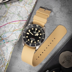 May include: A Tudor wristwatch with a black face and beige strap lies on a map. The watch has a black bezel with white markings and a date display. The strap is beige and has a silver buckle. A knife and a coin are also visible. The watch is a Tudor.