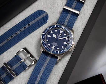 May include: A stainless steel wristwatch with a blue dial and a blue and gray striped nylon strap. The watch has a rotating bezel and is displayed with additional straps and a black box. The brand name "TUDOR" is visible.